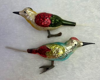 May include: Two vintage glass bird ornaments. One bird is yellow, red, and green, the other is blue, red, and yellow. Both have black beaks, metal clips, and feather tails. These ornaments are likely for a Christmas tree.