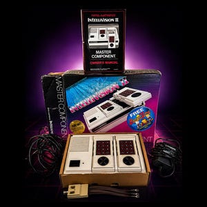 May include: Vintage Intellivision II video game console set. The box set includes the console, two controllers, a power supply, and the owner's manual. The box features the Intellivision II logo and the words "Master Component". The controllers have a white base with red number pads.