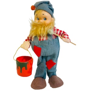 May include: A plush toy gnome wearing a blue hat, red and white checkered shirt, and blue overalls. The gnome is holding a paintbrush and a red paint can.