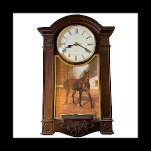 May include: A dark brown wooden clock with a round clock face featuring Roman numerals and gold trim. Below the clock face is a painting of a brown horse in a paddock. The clock has decorative carvings and a quartz movement.