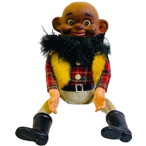May include: A vintage black-faced doll with a black beard, wearing a red and black plaid shirt, a yellow vest, and black boots.