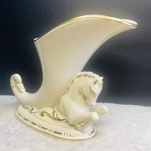May include: A white ceramic vase with a gold trim shaped like a horse's head and neck. The vase is decorated with gold accents and has a curved spout.