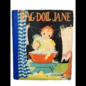 May include: A vintage hardcover book titled RAG-DOLL JANE with a black cover featuring an illustration of a doll with orange hair in an orange washbasin filled with soap bubbles. A blue and white checkered curtain hangs on the left side.