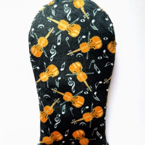 May include: A black golf club head cover with a pattern of orange violins and musical notes. The cover has a soft, plush texture and a drawstring closure. The design is repeated across the surface.