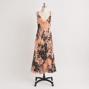 May include: A sleeveless midi dress featuring a peach-colored base with a black and light pink floral pattern. The garment has a draped cowl neckline, wide shoulder straps, and a flared skirt silhouette, displayed on a black rolling dress form.