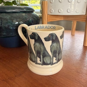 May include: Cream-colored ceramic mug featuring illustrations of black Labrador Retrievers. The word "LABRADOR" is printed in blue on the rim. The mug has a curved handle and a rounded base, perfect for coffee or tea.