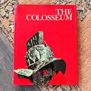 May include: A hardcover book with a bright red cover featuring the white text THE COLOSSEUM at the top. Below the text is a detailed illustration of a metallic Roman gladiator helmet with intricate relief carvings and a wide brim.