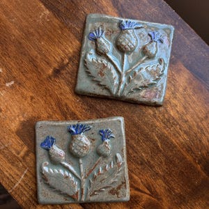 May include: Two square ceramic tiles featuring a raised relief design of thistle plants. The tiles have a sage green glaze with brown accents and vibrant blue thistle flower heads. Each tile displays a botanical motif with textured leaves and stems.
