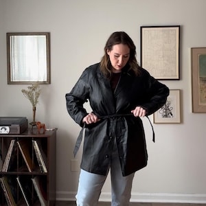 May include: A black leather jacket with a belt and a button closure. The jacket is worn over a light blue denim shirt.