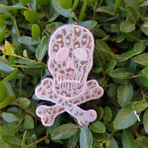 May include: A pink and brown skull and crossbones patch with a leopard print design. The patch is embroidered and has a black backing.
