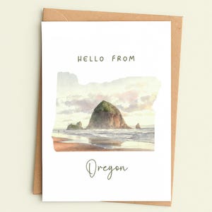 May include: A greeting card with a watercolor painting of a coastal landscape. The card features a large rock formation in the ocean, with the words "HELLO FROM" and "Oregon" printed on the front. The card is inside a brown envelope.