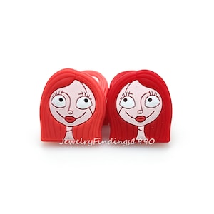 May include: Two red silicone hair ties featuring a cartoon image of a girl with red hair and a white face. The girl has large eyes, a small nose, and a red mouth.