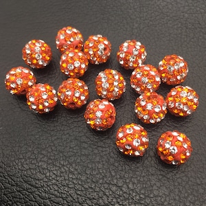 May include: A group of 16 orange and clear rhinestone beads. The beads are round and have a sparkly finish.