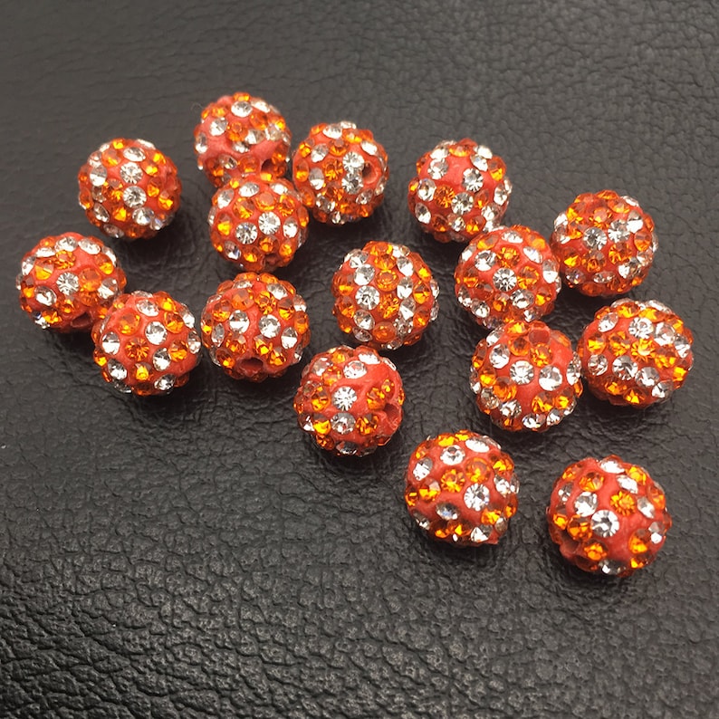 May include: A group of 16 orange and clear rhinestone beads. The beads are round and have a sparkly finish.