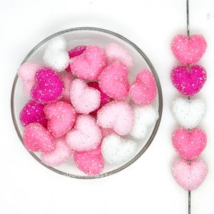 Pode incluir: Uma tigela de vidro transparente cheia de decorações em forma de coração rosa, branco e fúcsia. Alguns corações são enfiados em um fio. Os corações são cobertos com glitter, criando um efeito brilhante. Em um fundo branco.