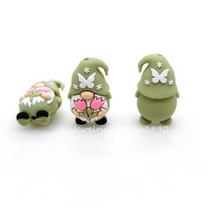 May include: Three green gnome figurines with white butterfly and star details. One gnome is lying on its side, while the other two are standing. One of the standing gnomes holds pink tulips. The text "JewelryFindings1990" is visible.