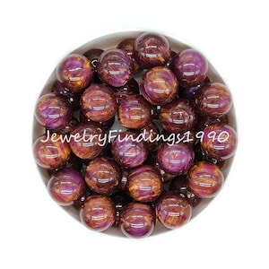 May include: A bowl filled with 20 large, round, purple and gold acrylic beads. The beads have a swirled, marbled effect.