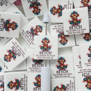 May include: A collection of square white tiles featuring a colourful illustration of a monkey sitting in a lotus position, wearing a cap and smoking. Below the monkey, the text reads BITCH You breathe too much Shut the fuck up. A metal tool is visible.