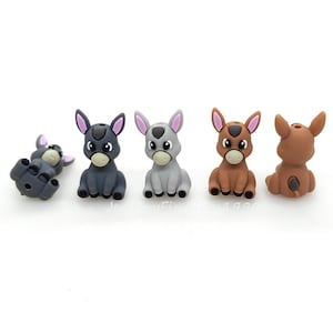 May include: A collection of five donkey figurines in various poses and colors. The donkeys are in shades of gray, brown, and tan, with pink or purple accents on their ears. One donkey is lying on its side, while the others are sitting upright.