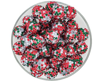 May include: A collection of round, decorative beads in a clear glass bowl. The beads are covered in red, green, and silver rhinestones. The beads are likely for jewelry making or other craft projects.
