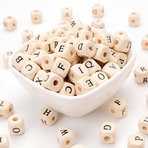 May include: A collection of small, natural wood letter beads, each cube-shaped with a drilled hole. The beads are scattered around and inside a white ceramic bowl. Each bead has a different black letter printed on its faces.
