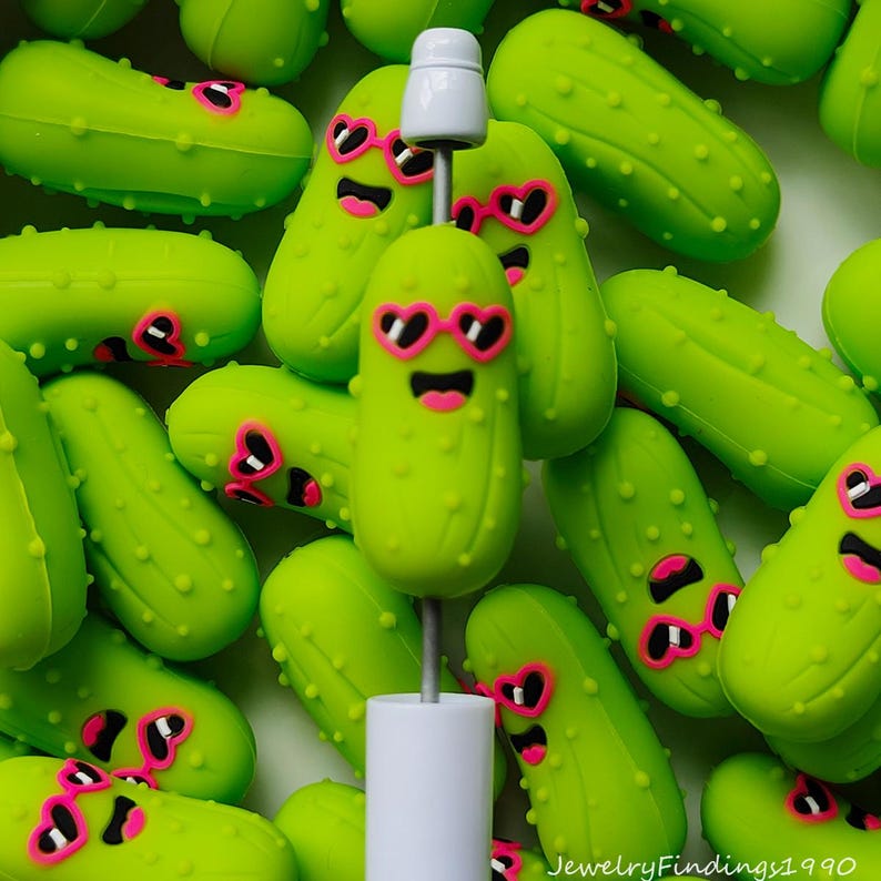 Puede incluir: Una colecci&oacute;n de amuletos en forma de pepinillo verde brillante, algunos con gafas de sol rosas en forma de coraz&oacute;n y caras sonrientes. Un tap&oacute;n de botella blanco con un amuleto de pepinillo est&aacute; en el centro.