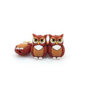 May include: Three brown owl-shaped silicone beads with yellow eyes and white accents. The owls are facing forward with their wings spread out.