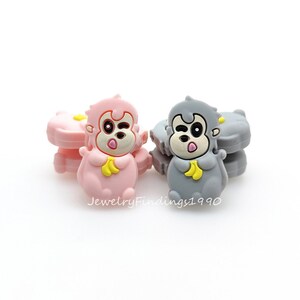May include: Two silicone monkey charms, one pink and one grey, with a yellow banana in each hand. The monkey charms have a cartoon-like design with big eyes and a winking expression.