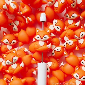 May include: A collection of small orange and white fox-shaped beads with closed eyes, featuring one fox bead mounted on a thin metal rod with a white handle. The fox beads have a smooth finish and a curled tail design, with white accents on the chest.