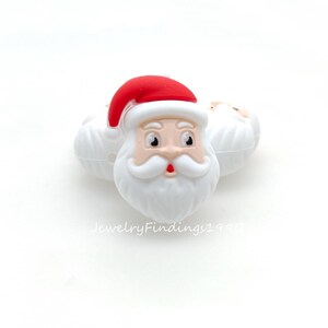 May include: White Santa Claus head-shaped ornaments with red hats and white beards. The ornaments have a smiling face with a red mouth. The text "JewelryFindings1990" is visible on the bottom.