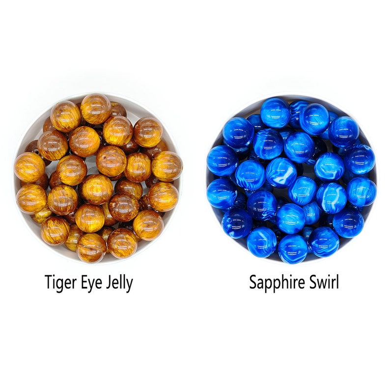 May include: Two bowls of colorful beads. The bowl on the left contains brown and gold beads with a tiger eye pattern. The bowl on the right contains blue and white swirl beads. The text "Tiger Eye Jelly" is below the left bowl and "Sapphire Swirl" is below the right bowl.