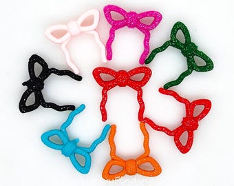 May include: A collection of colorful bow-shaped embellishments in various colors, including pink, red, green, blue, orange, and black. Each bow has a glittery texture and is arranged on a white background. These decorative bows are suitable for crafts and jewelry making.