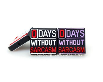 May include: Two black square shoe charms with white text that reads "0 DAYS WITHOUT SARCASM". One charm has a red "0" and the other has a purple "0".