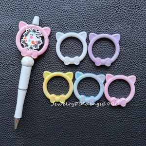 May include: Six colorful cat-shaped rings with bows. The rings are made of plastic and have a glossy finish. The rings are in various colors, including pink, purple, white, yellow, and blue.