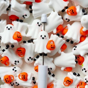May include: A collection of white ghost-shaped charms, each featuring a small orange jack-o'-lantern bucket. One ghost is mounted on a white-handled metal tool. The ghosts have black eyes, a small mouth, and pink cheeks, with black feet at the base.