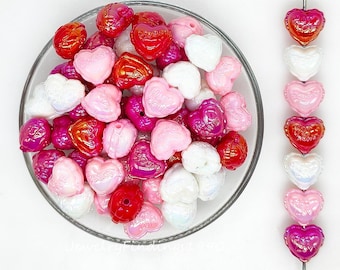 May include: A collection of heart-shaped beads in a glass bowl and strung on a wire. The beads are in shades of red, pink, white, and purple. Some beads have the words "Sweet Love" or "Hot Mom" on them. The beads are for jewelry making.