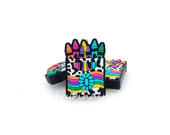 May include: A colorful box of crayons with a black and white cow print design. The crayons are arranged in a box with a rainbow stripe and a blue flower.