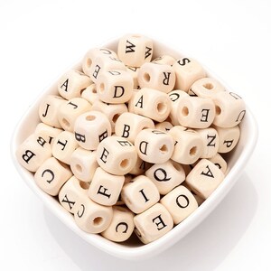 May include: A collection of wooden alphabet beads in a white ceramic bowl. Each cube-shaped bead has a letter printed in black on each side. The beads are a natural wood color.
