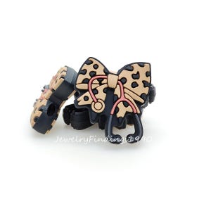 以下が含まれることがあります： 黒とタン色の靴チャームのペア。1つのチャームは、ヒョウ柄のデザインとピンクの聴診器が付いたリボンが特徴です。もう1つのチャームはヒョウ柄のデザインです。