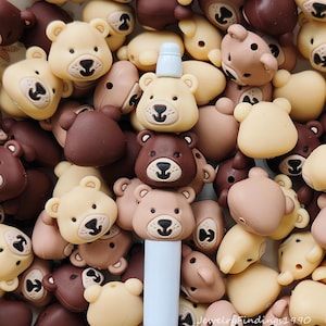 May include: A collection of bear-shaped beads in various shades of brown and tan. The beads have black eyes and noses, with a few featuring a small, white cylindrical object on top. The beads are scattered, creating a playful and whimsical aesthetic.