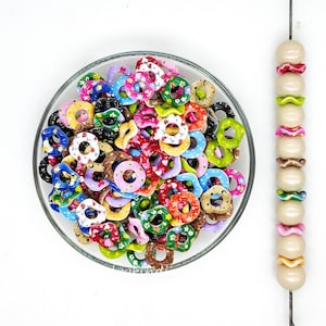 Peut inclure: Un ensemble de perles colorées en forme de donut dans un bol en verre transparent. Les perles sont de différentes couleurs, notamment rose, vert, bleu et jaune, avec des motifs floraux et à motifs. Une rangée de perles crème avec des accents décoratifs est à droite.