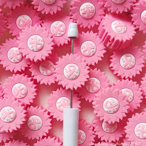 May include: A collection of pink sunflower-shaped beads featuring a white centre with a pink ribbon emblem and small star accents. One bead is mounted on a thin metal rod with a white handle. The text JewelryFindings1990 is visible at the bottom.