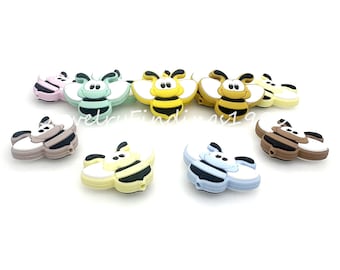 May include: A set of eight silicone baby teethers shaped like bees in various colours, including yellow, green, blue, brown, and pink. Each bee has black stripes and a friendly face.