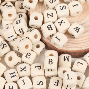 May include: A collection of small, natural wood letter beads. Each cube-shaped bead has a black letter printed on it, with a hole drilled through the center. The letters are a mix of the alphabet.