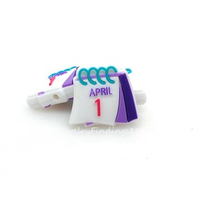 May include: A calendar charm with the date April 1st. The charm is white with purple and teal accents. The calendar is open to the first day of April. The charm is on a white background.