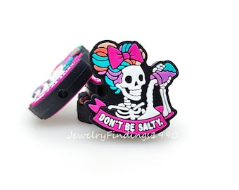 May include: A stack of black and pink charms with a skeleton design. The skeleton has a pink bow, teal hair, and is holding a purple drink. The text "DON'T BE SALTY" is on a pink banner. The charms are for jewelry making.