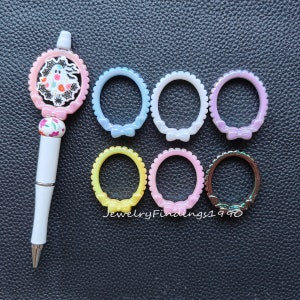 May include: Seven colorful plastic rings with a bow detail. The rings are in shades of blue, purple, yellow, and pink. The rings are arranged in a semi-circle on a black surface.  JewelryFindings1990 is printed on the surface.