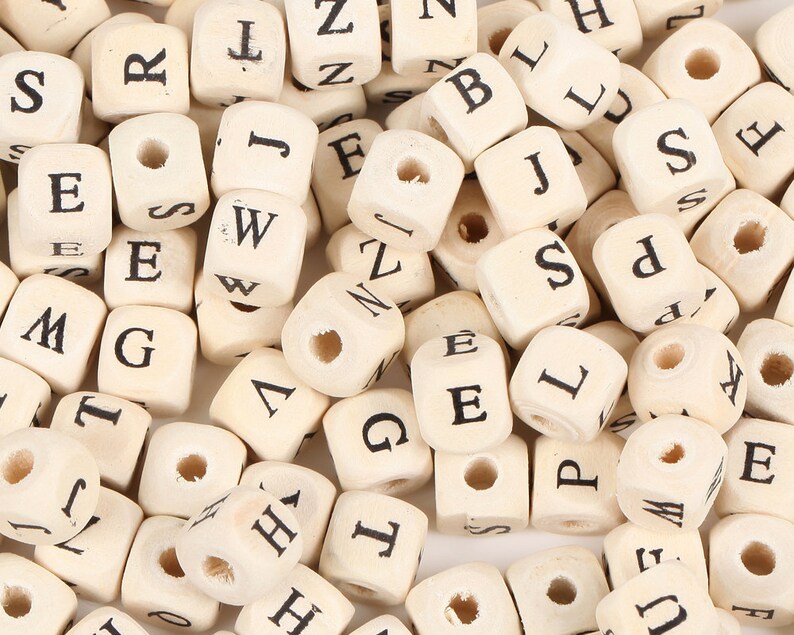 May include: A collection of small, natural wood letter beads. Each cube-shaped bead has a single letter printed in black. The beads are undrilled, with a hole running through the center, and are scattered across a white surface.