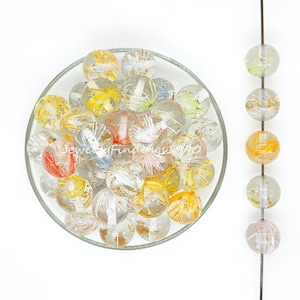 Może przedstawiać: Miska z przezroczystymi szklanymi koralikami z złotymi drobinkami i kolorowymi wtrąceniami. Korale są okrągłe i mają przezroczysty wygląd. Na drucie widoczny jest sznur takich samych koralików.