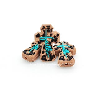 以下が含まれることがあります： 茶色と黒のヒョウ柄のクロス型ビーズ。ターコイズ色の筆記体で「Faith」の文字が書かれています。ビーズは明るい茶色で光沢があります。白い背景にビーズがまとめられています。